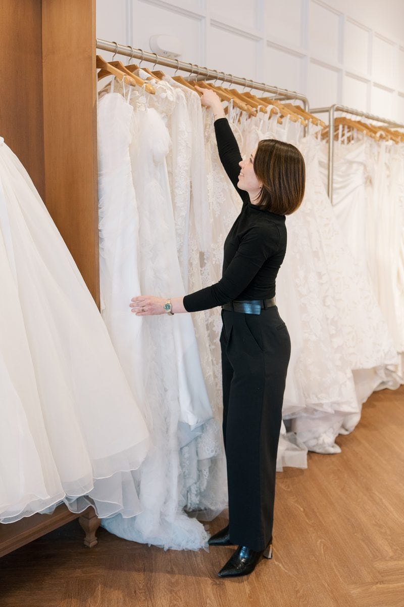 stylist dressed in all black placing a gown back on the rack full of other white bridal gowns