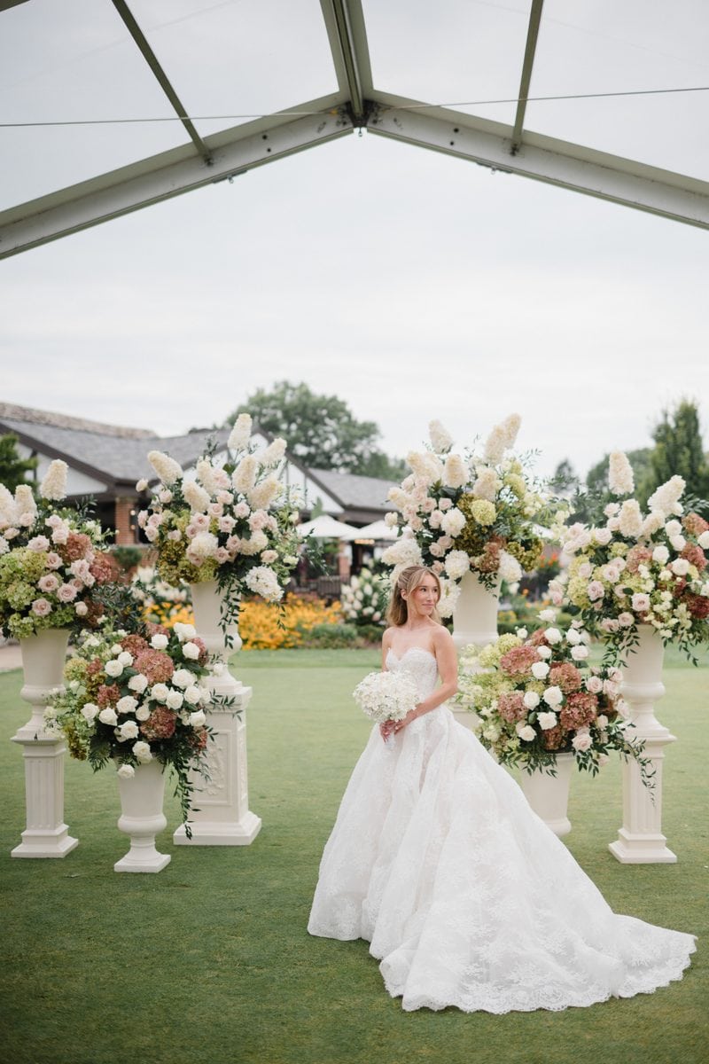 a bride in a lace ball gown at her outdoor ceremony venue surrounded by white tall vases with white, green, and pink florals